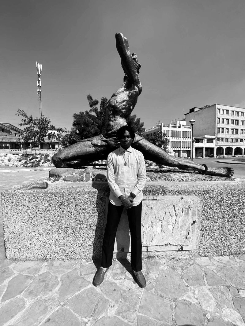 Roth$child standing in front of the Nèg Mawon statue in Port-au-Prince under a clear sky.