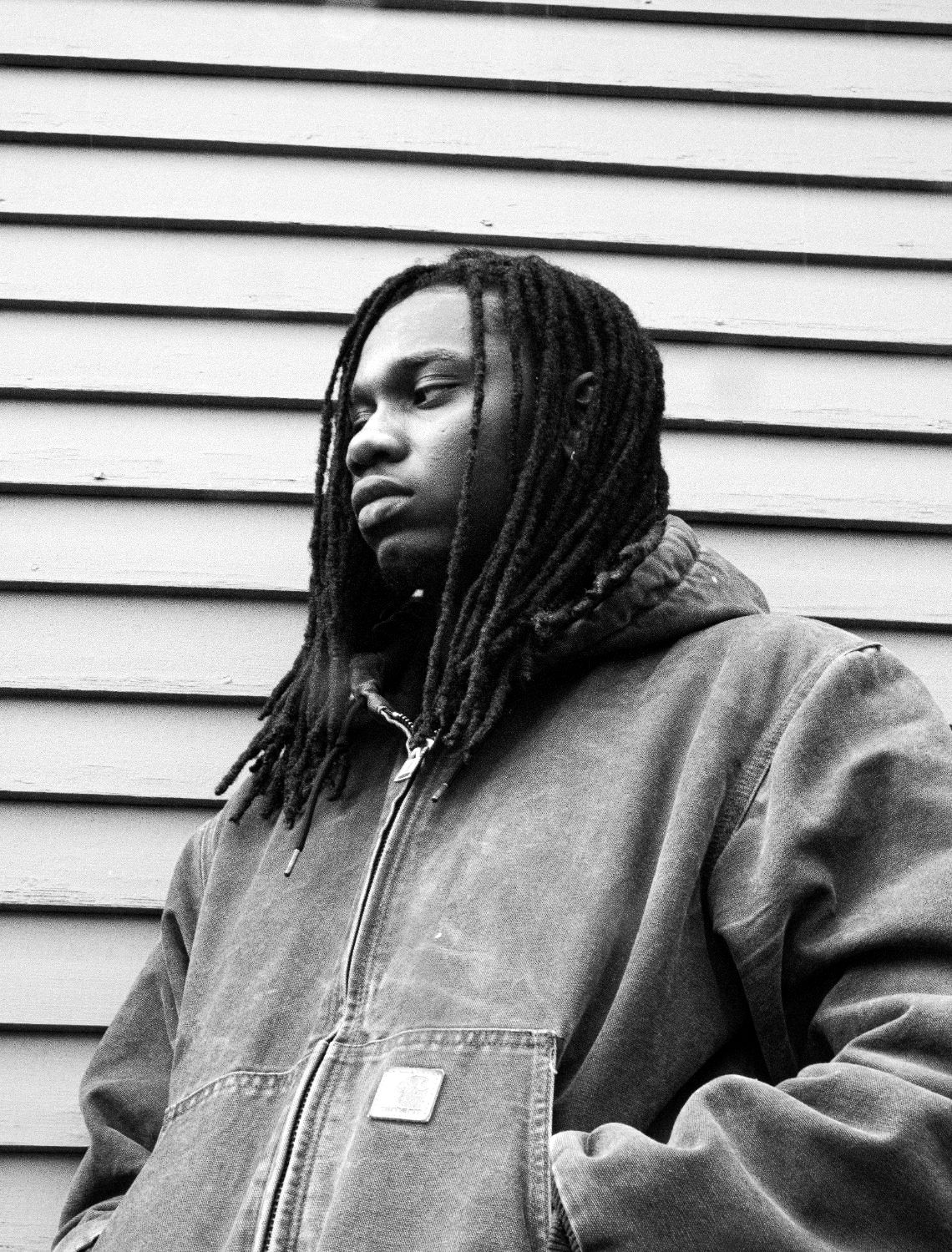 Roth$child in a worn canvas work jacket, locs falling past the shoulders, against pale clapboard siding.